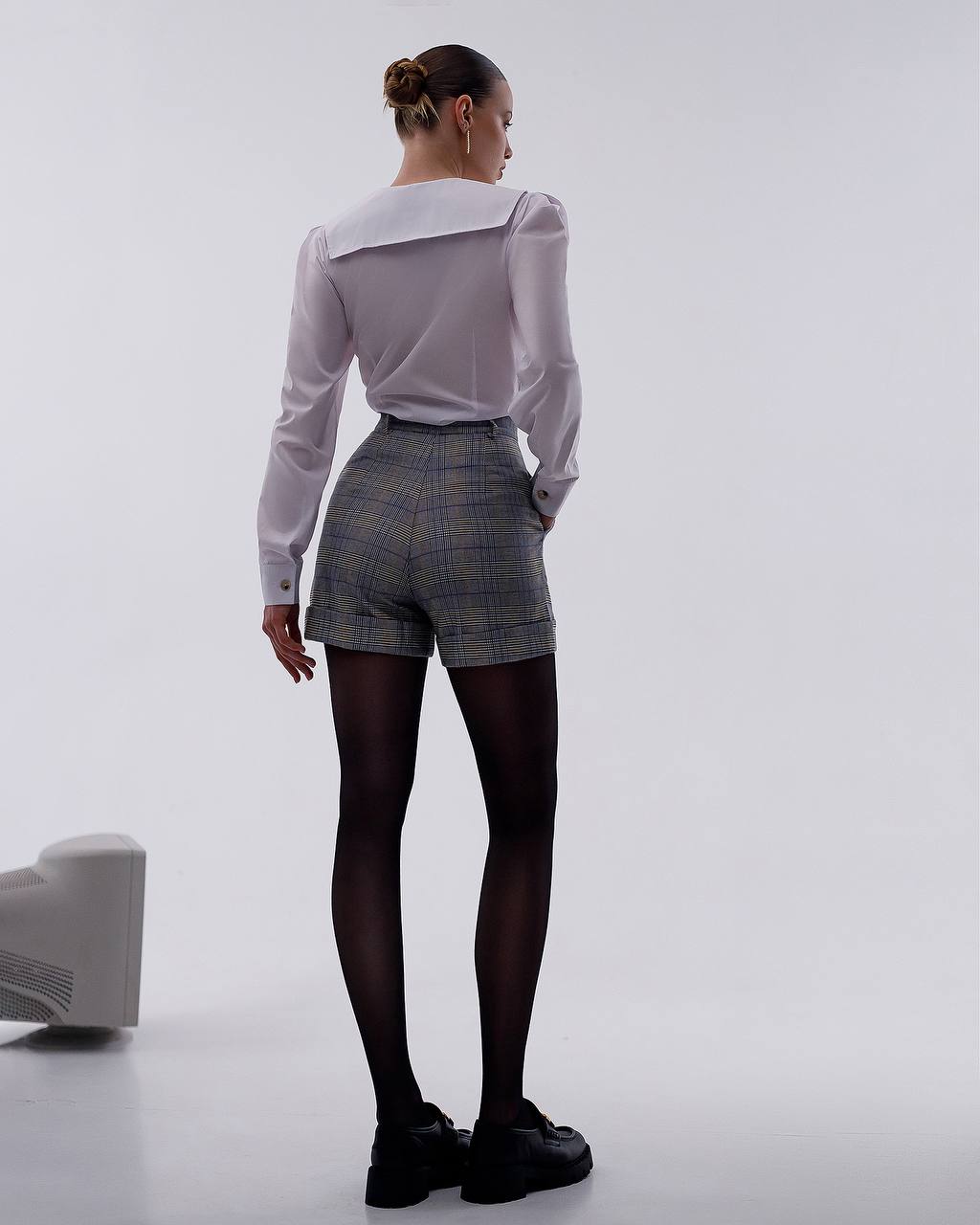 Person wearing a white blouse, plaid shorts, and black tights on a plain background