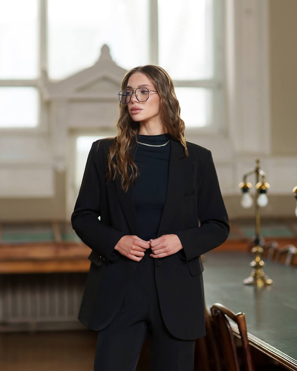 Woman wearing a black suit standing in an elegant room with large windows.