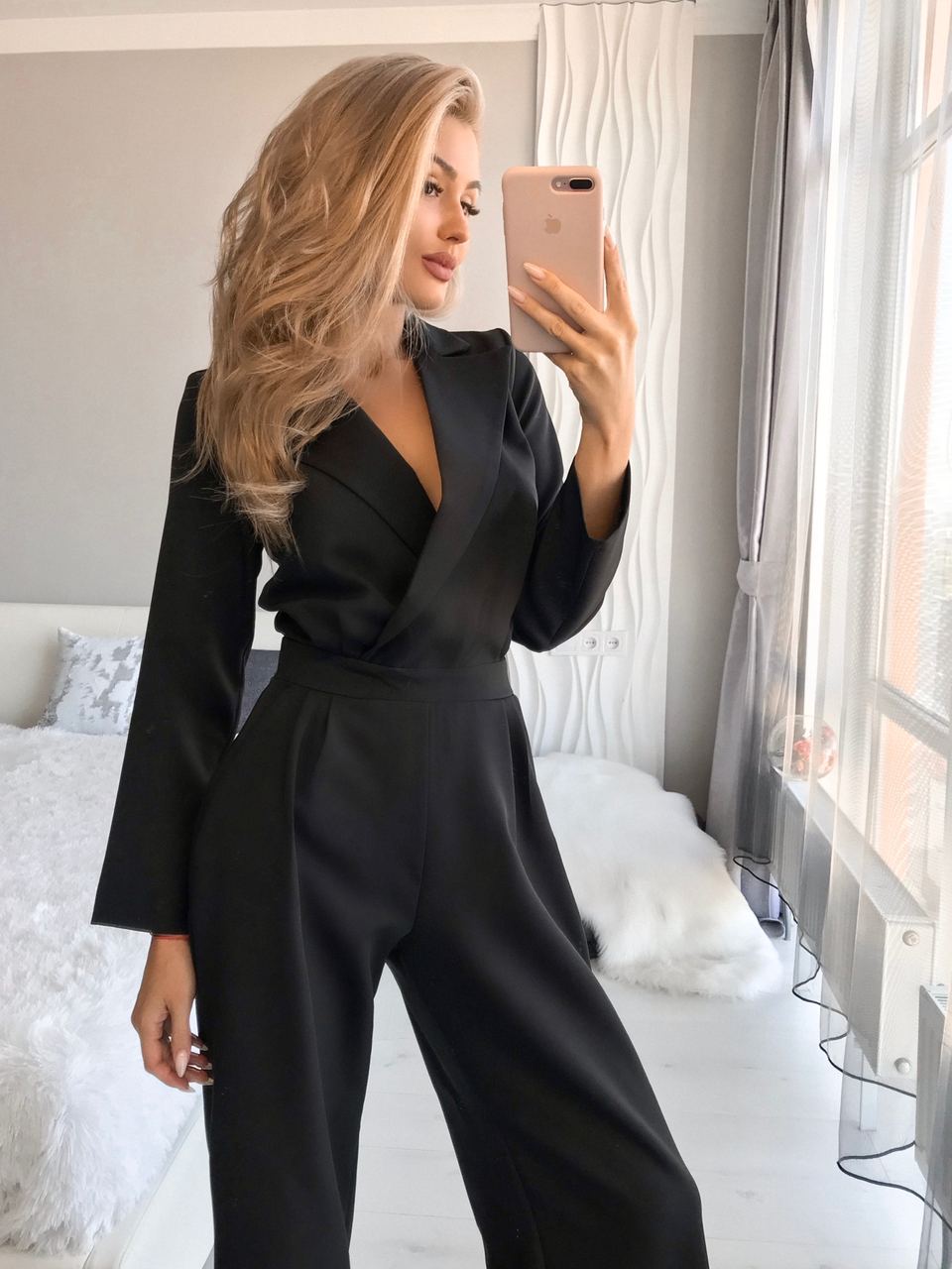 Woman taking a mirror selfie wearing a black jumpsuit in a bright room.