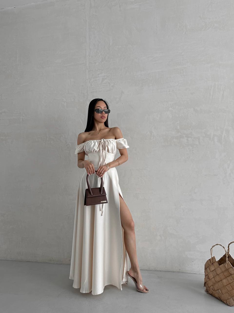 Woman in a white off-shoulder dress with a high slit, holding a brown handbag against a plain background.