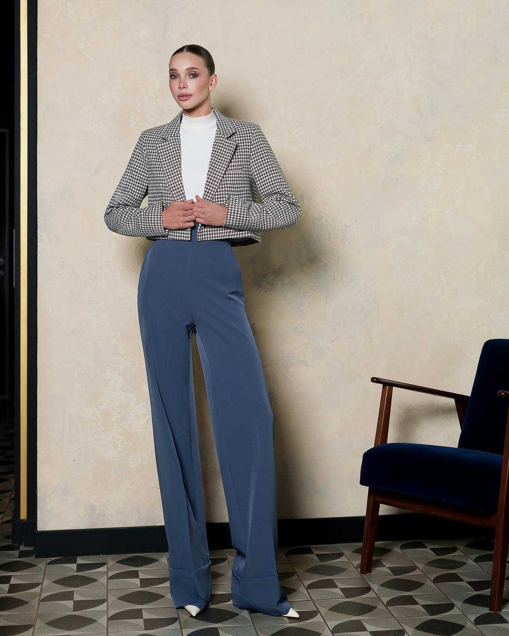 Woman in a checkered blazer and blue pants standing in a room with a chair and patterned floor.