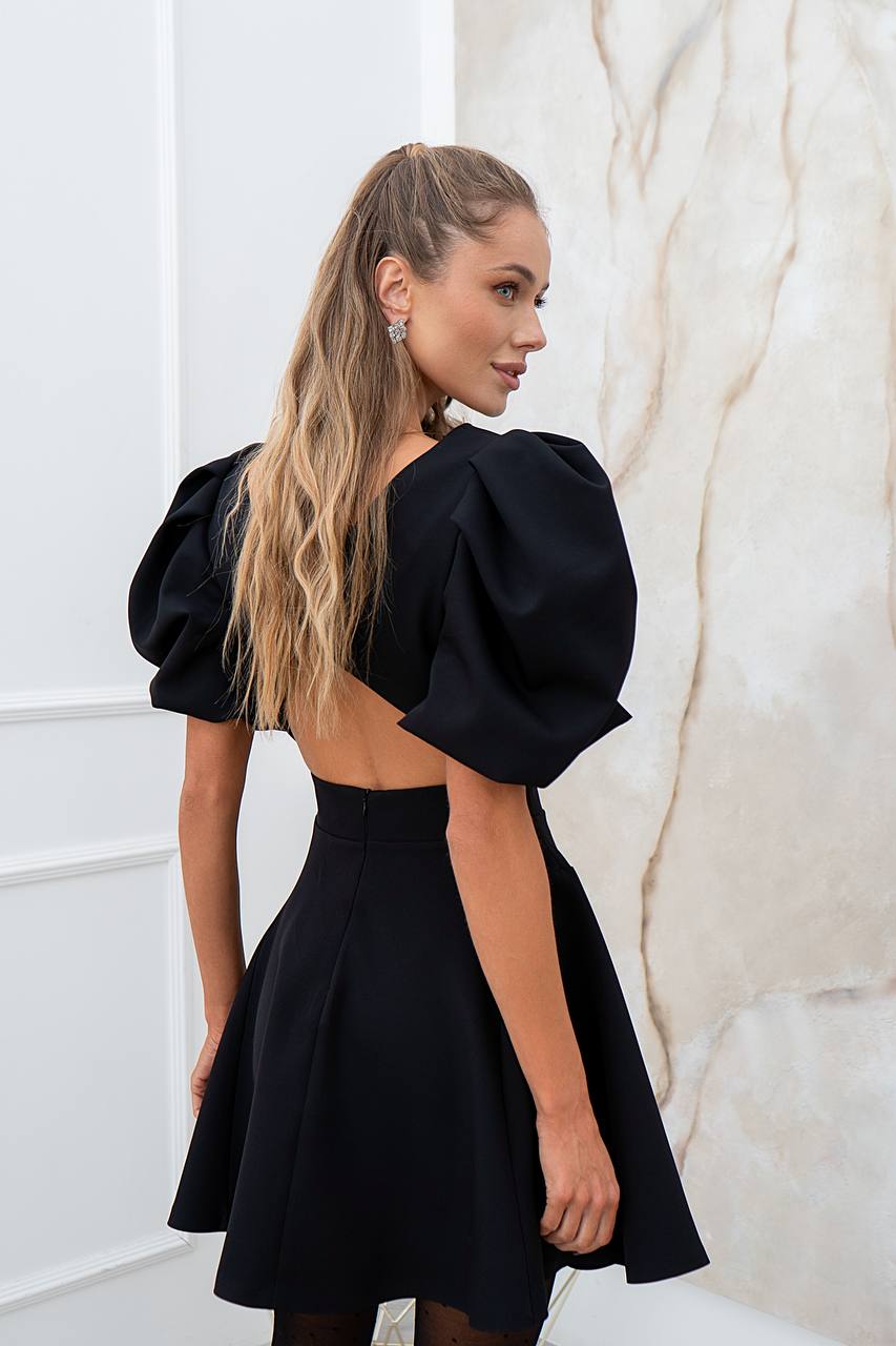Woman wearing a black dress with puffed sleeves against a white wall.
