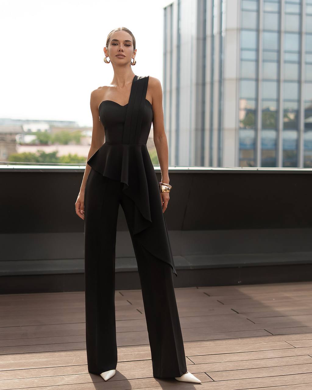 Woman wearing a black one-shoulder jumpsuit on a rooftop with cityscape background