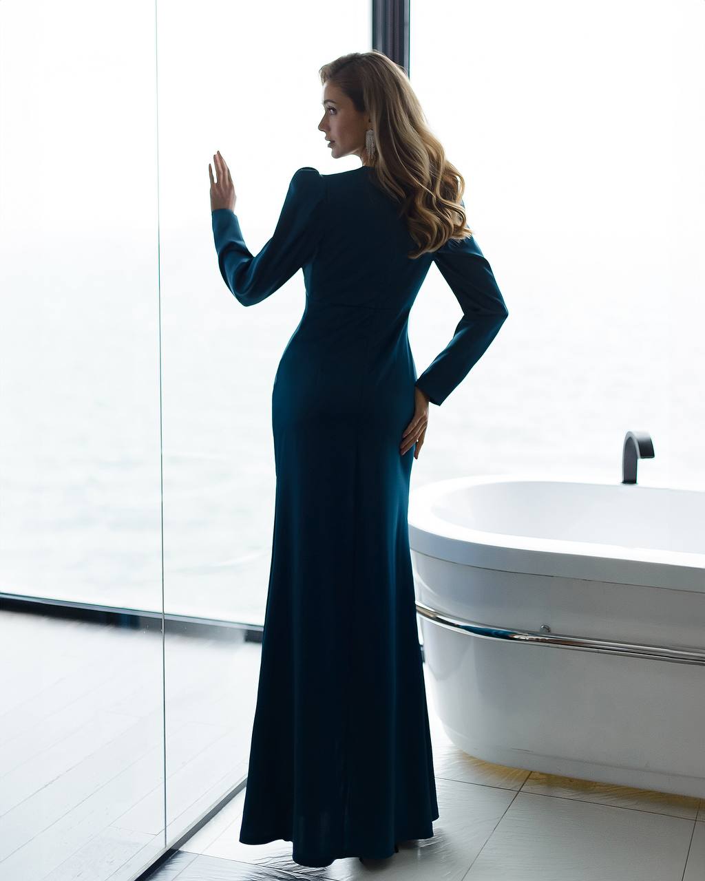 Woman in a teal dress standing by a bathtub in a modern bathroom.