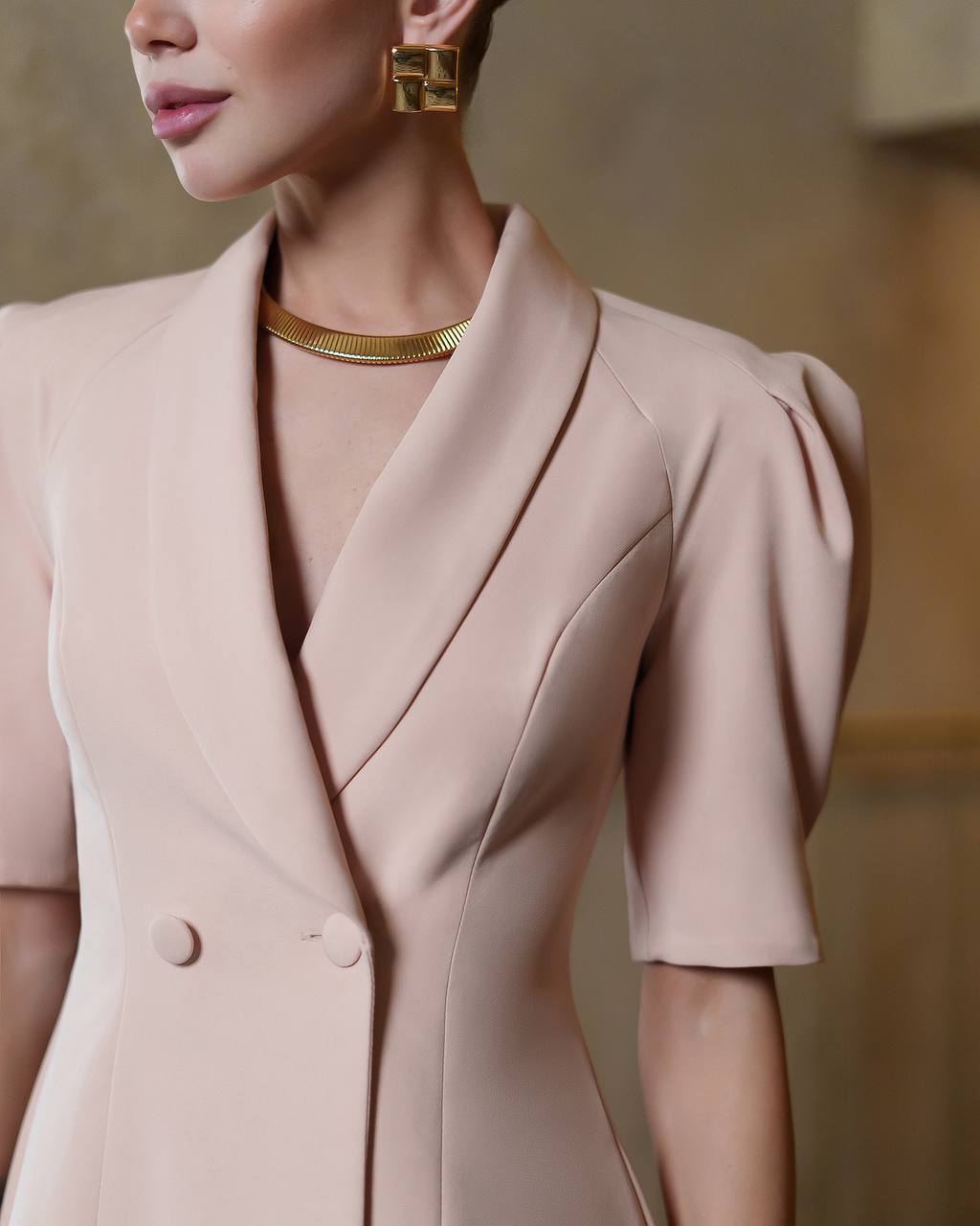 Close-up of a person wearing a light pink blazer with gold jewelry against a neutral background