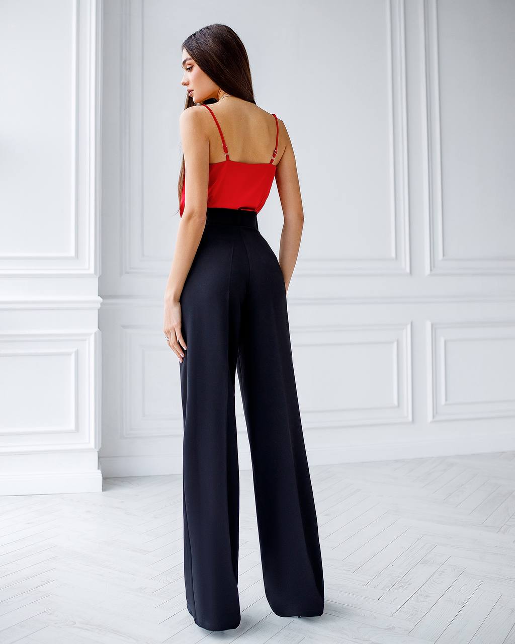 Woman wearing a red top and black high-waisted pants in a white room.