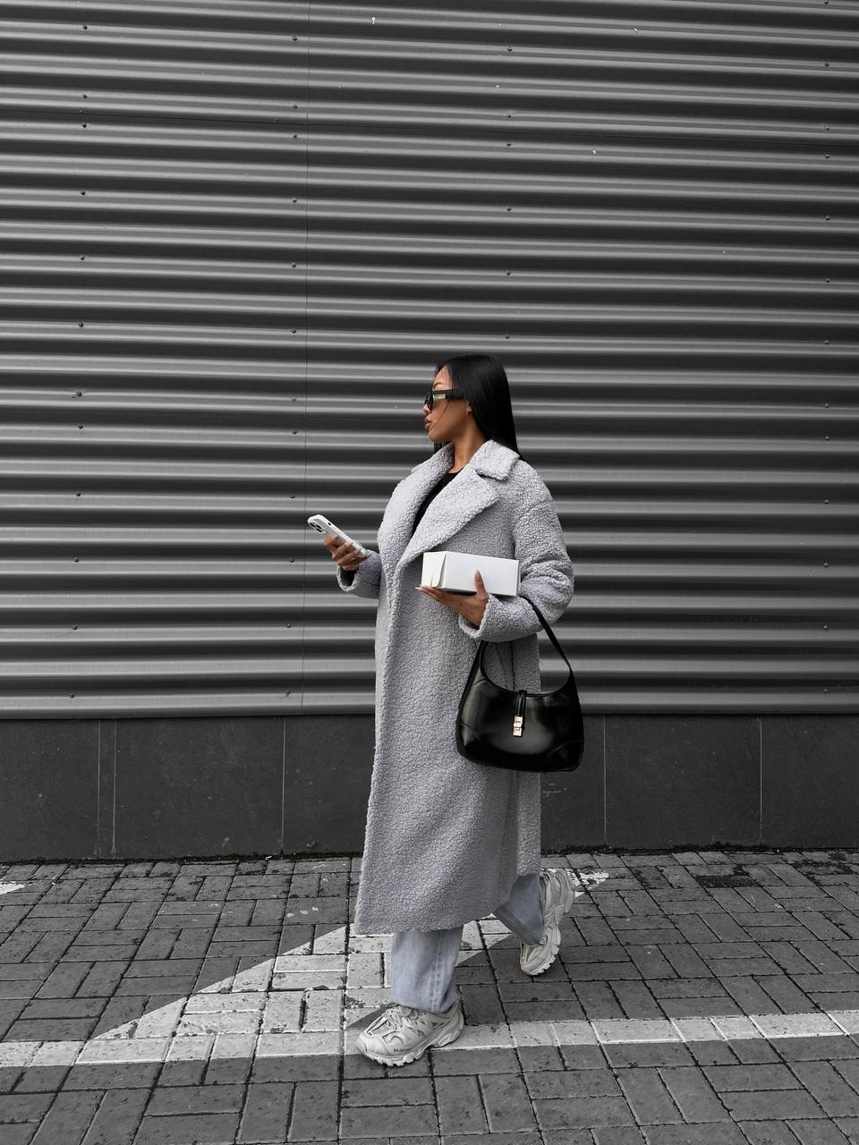 Woman in a gray coat standing against a corrugated metal wall.