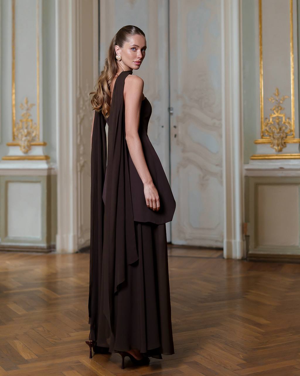 Woman in a long brown dress standing in an elegant room with ornate walls.