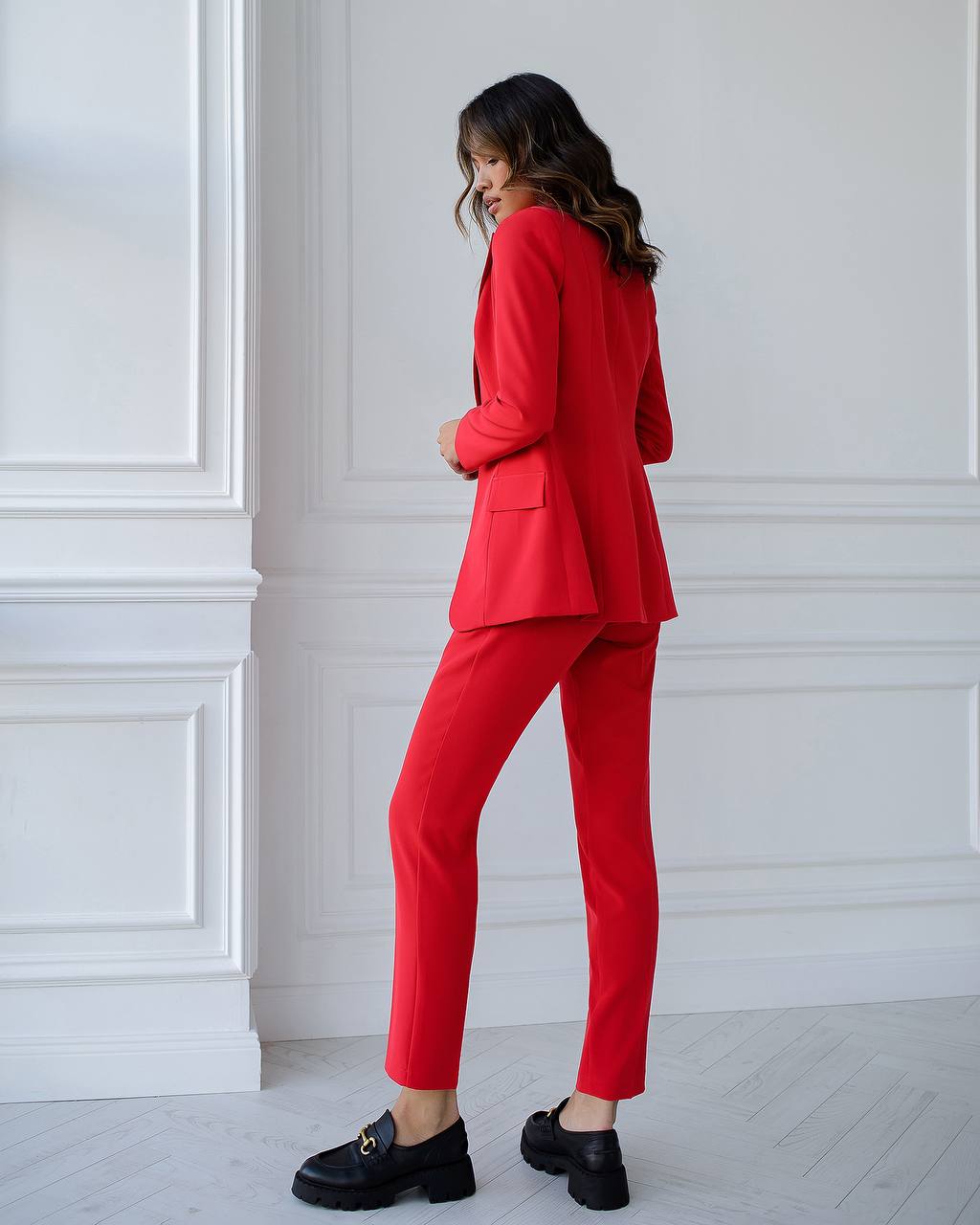 Woman wearing a red suit standing against a white paneled wall.