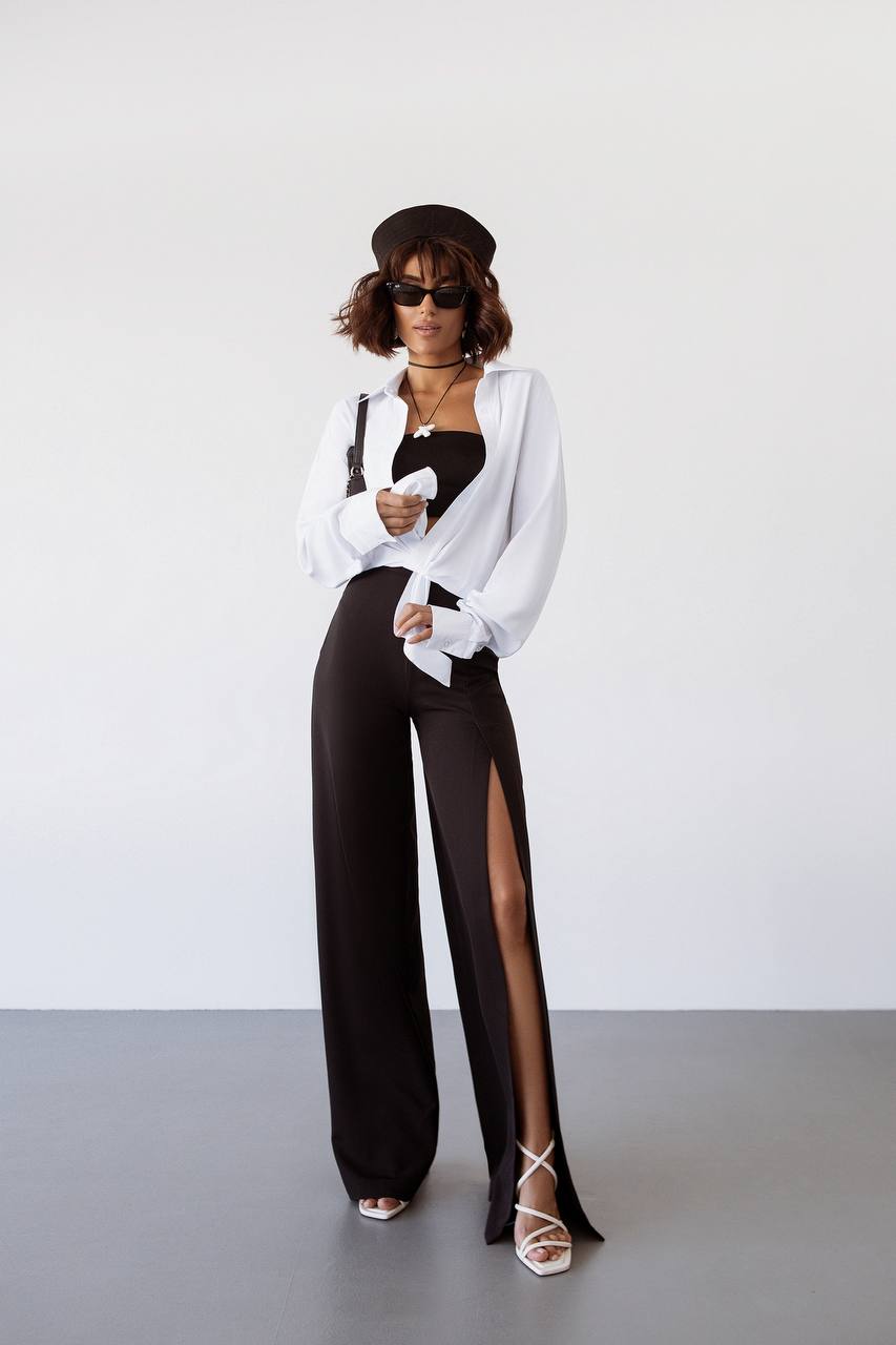 Woman wearing a white blouse, black pants with a high slit, and sunglasses on a plain background
