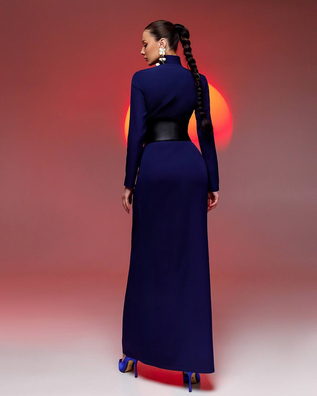 Woman in a long blue dress with a high collar and black belt, standing against a gradient red to pink background.