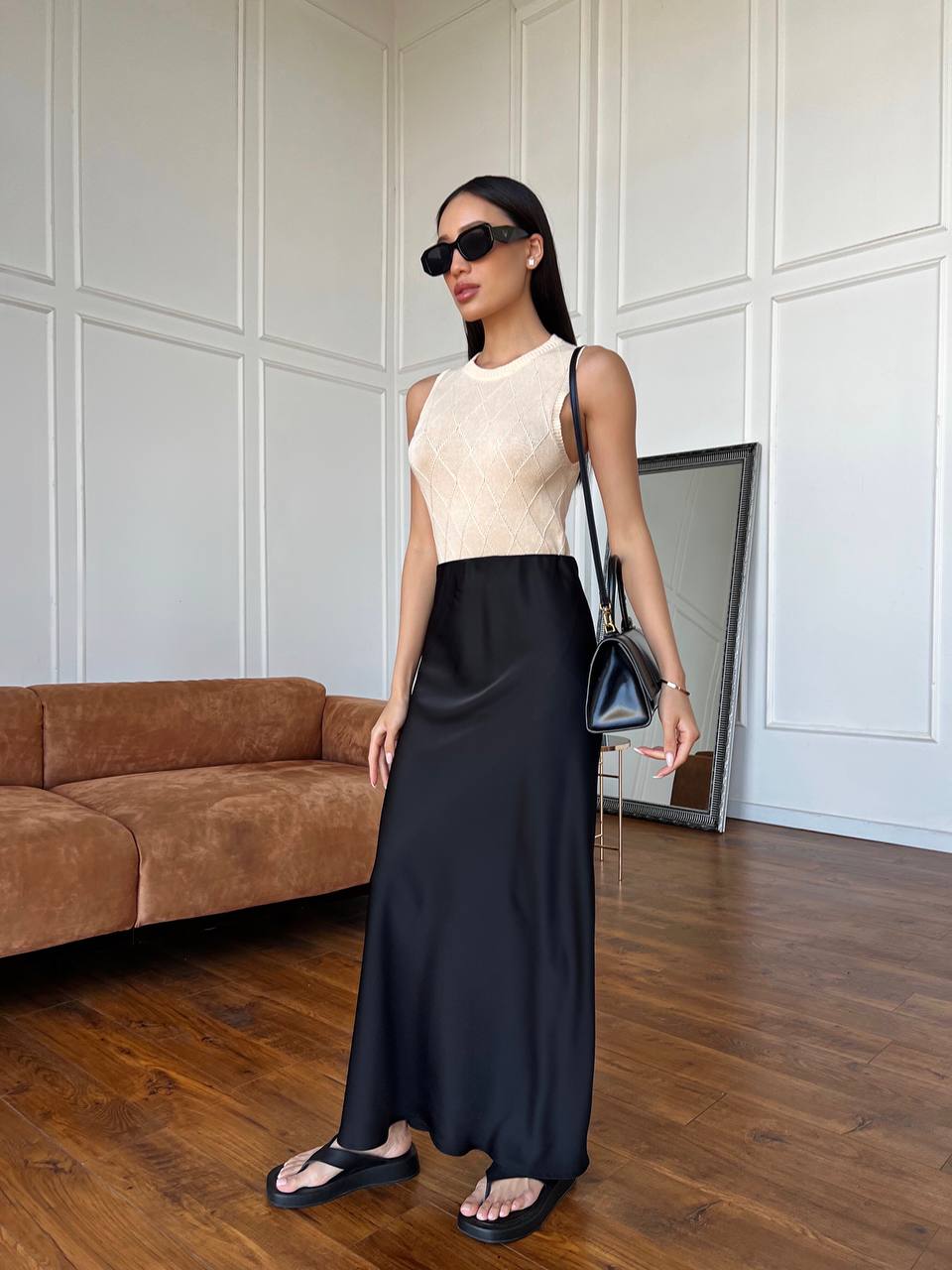Woman in a beige top and black skirt standing in a room with a brown couch and white paneled walls.