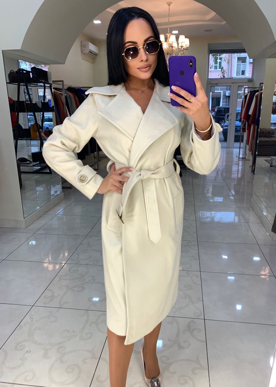 Woman taking a mirror selfie wearing a white trench coat in a modern interior setting.