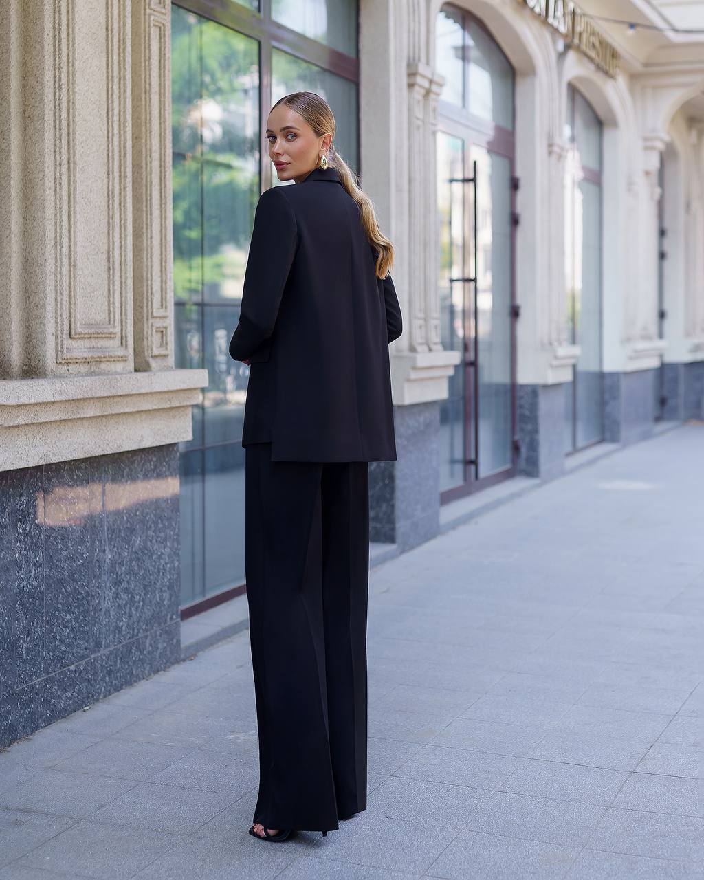 Woman in a black suit standing on a city street with classical architecture.