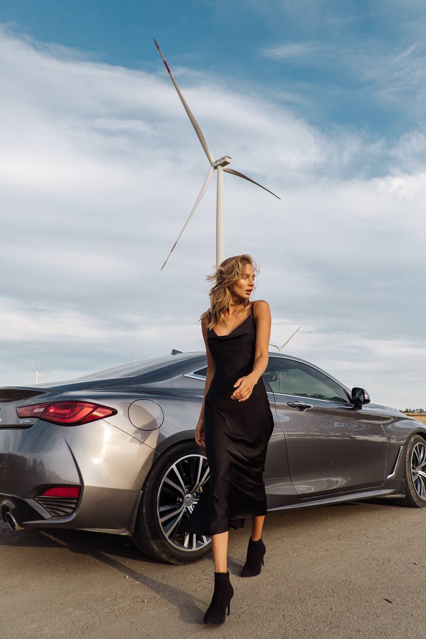 Woman in a black dress standing next to a silver car with a wind turbine in the background