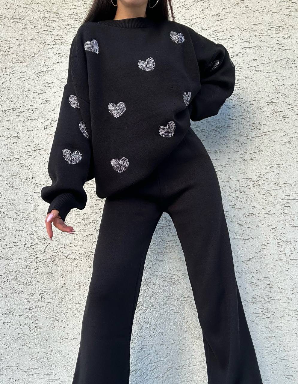 Person wearing a black sweater with heart patterns against a textured wall.