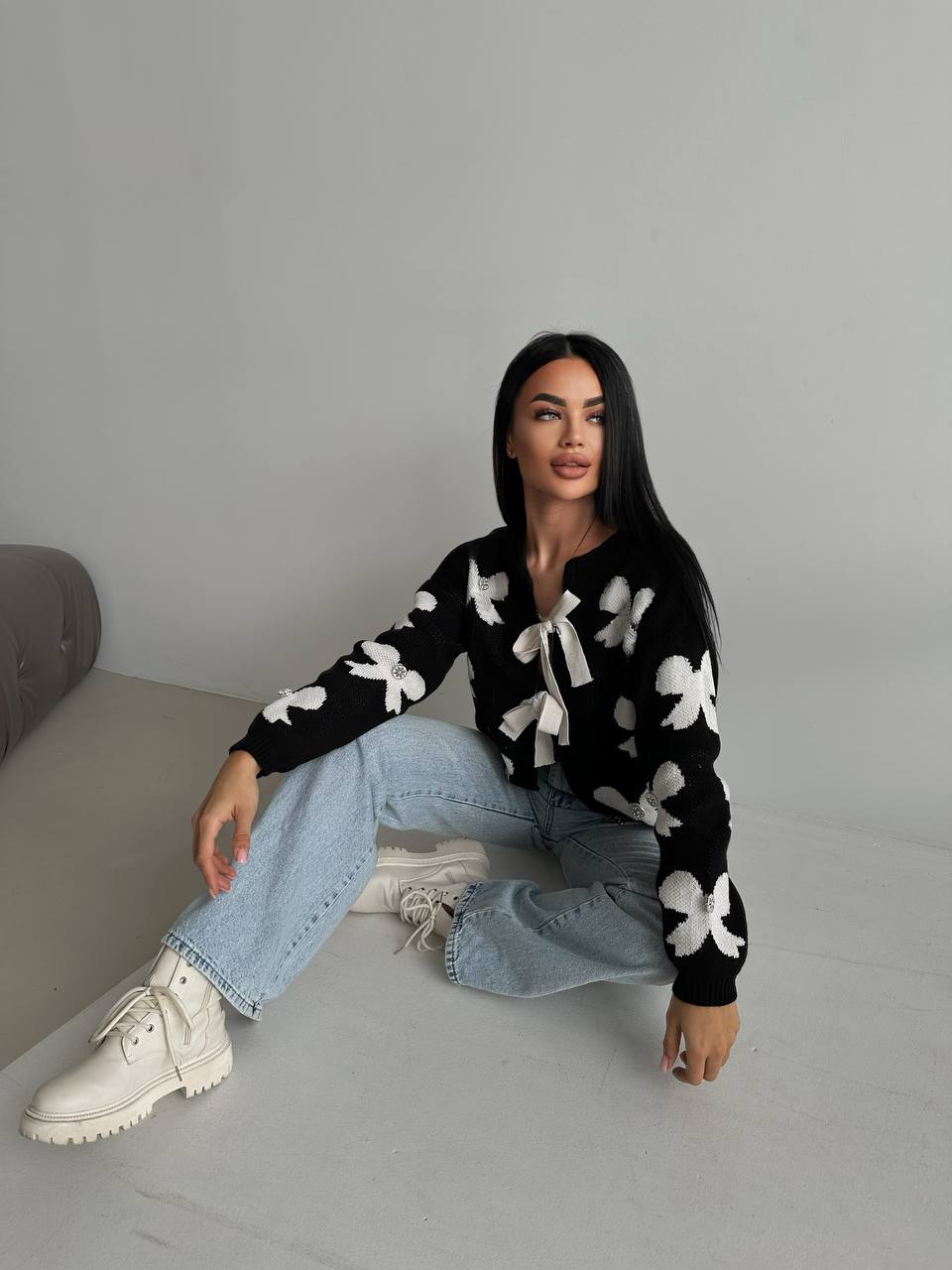 Woman wearing a black sweater with white patterns, light blue jeans, and white boots sitting on a gray surface.