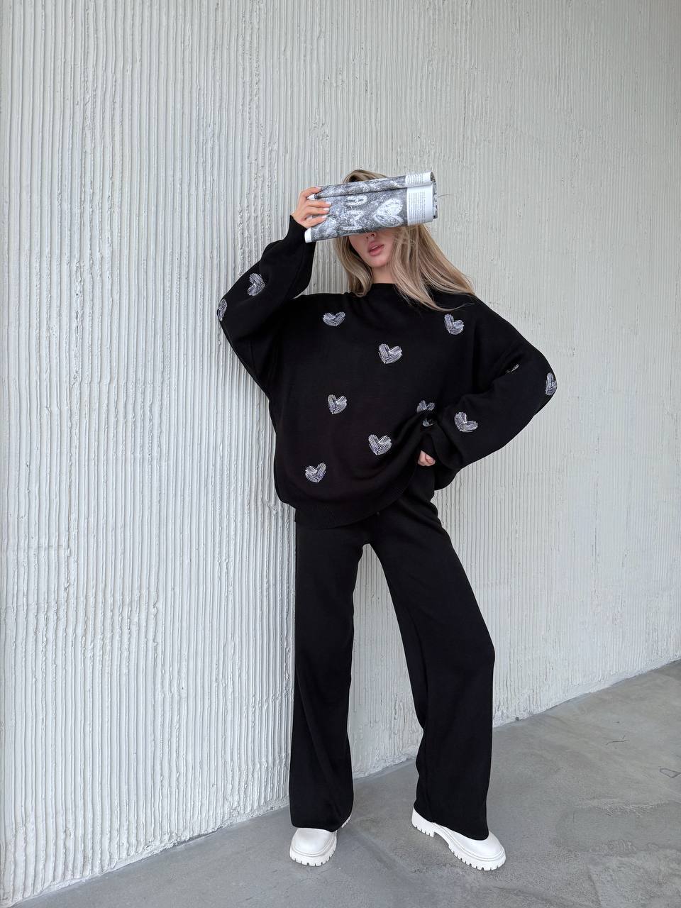 Person wearing a black outfit with heart patterns against a textured wall.