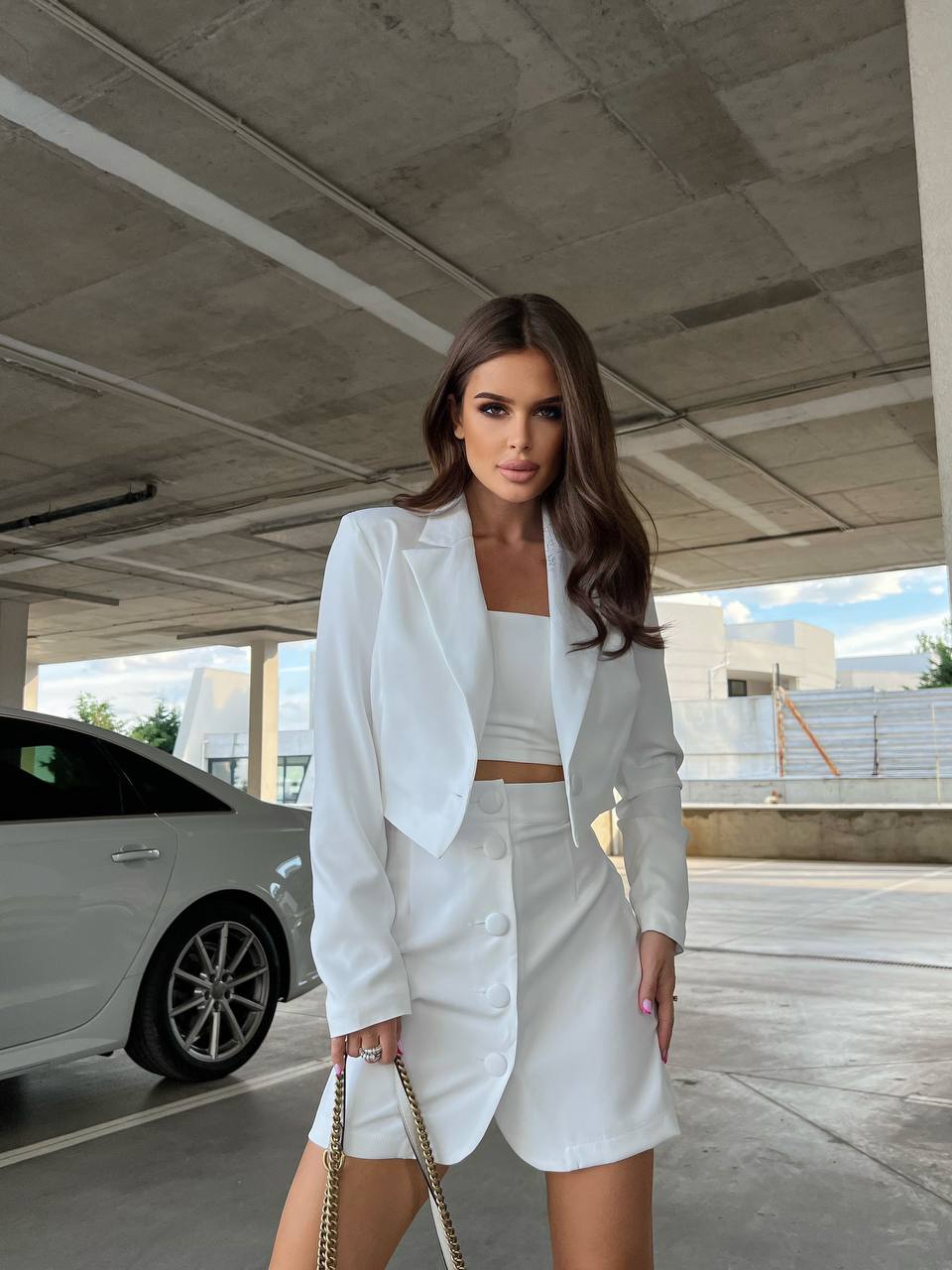 Woman in a white outfit standing in an outdoor setting with a car and building in the background.