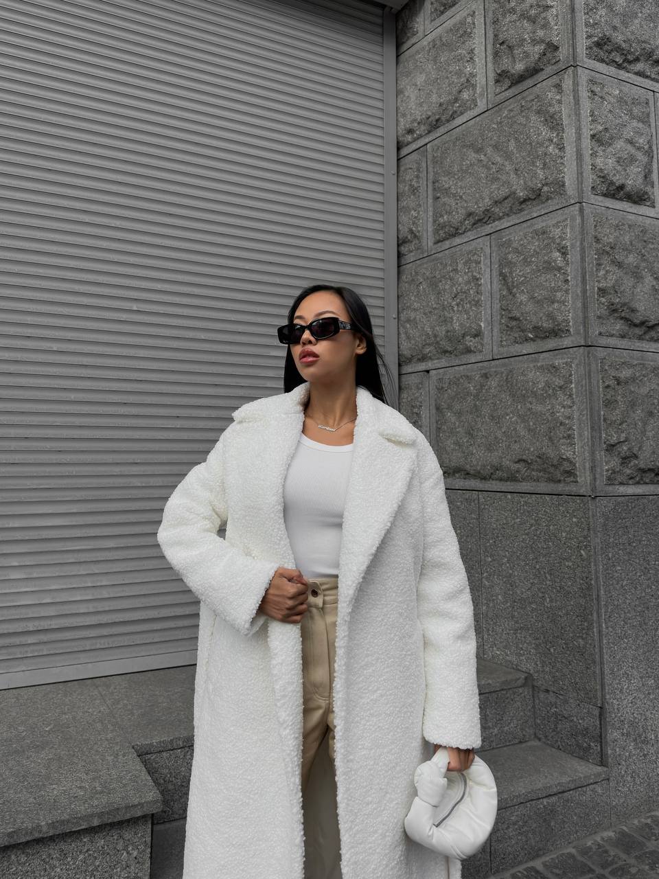 Woman wearing a white coat and sunglasses standing against a gray wall.