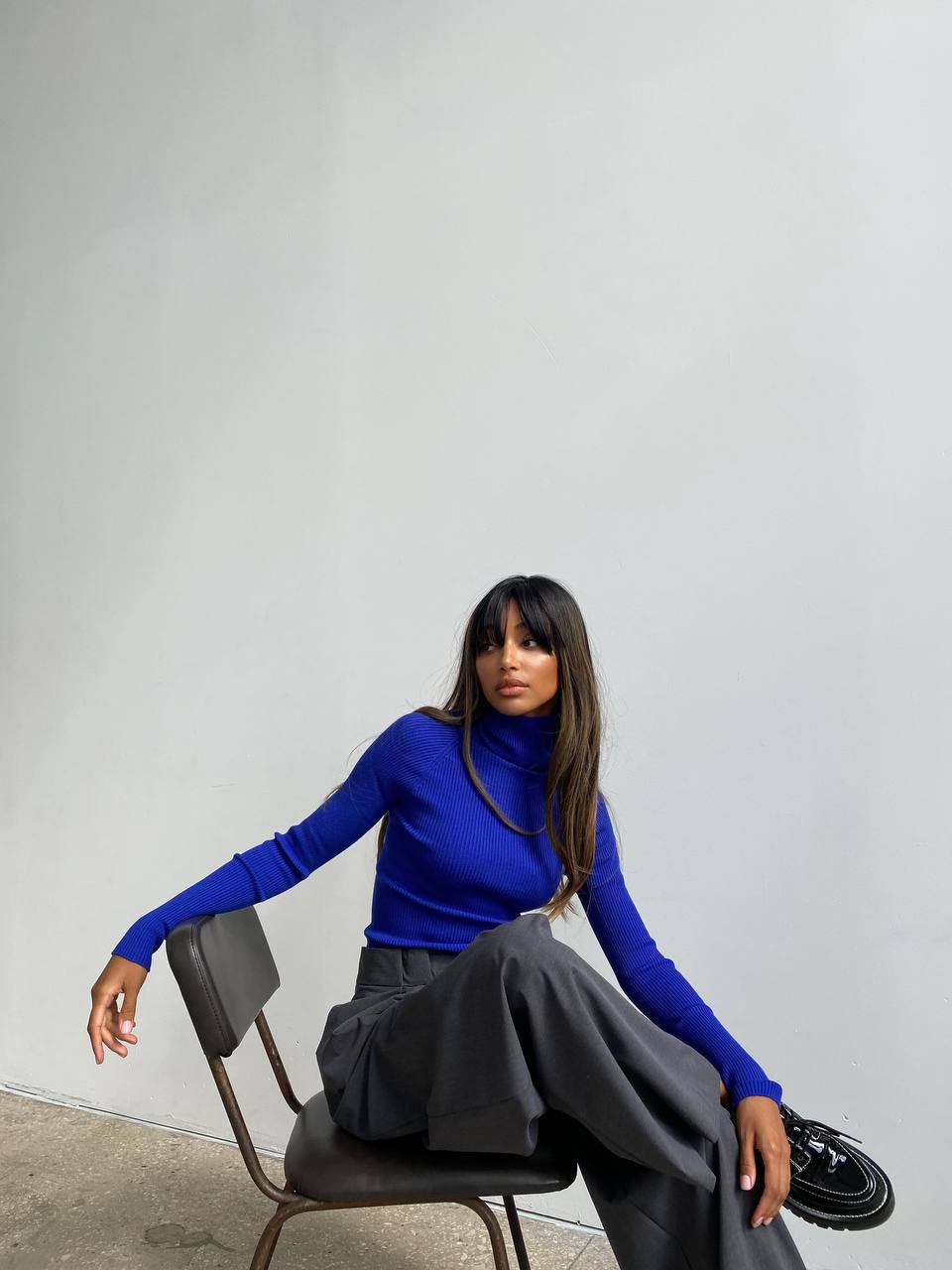 Person wearing a blue sweater and gray pants sitting on a chair against a plain background