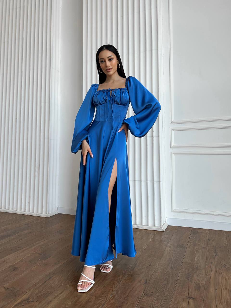 Woman wearing a blue dress with a high slit in a room with white paneled walls.