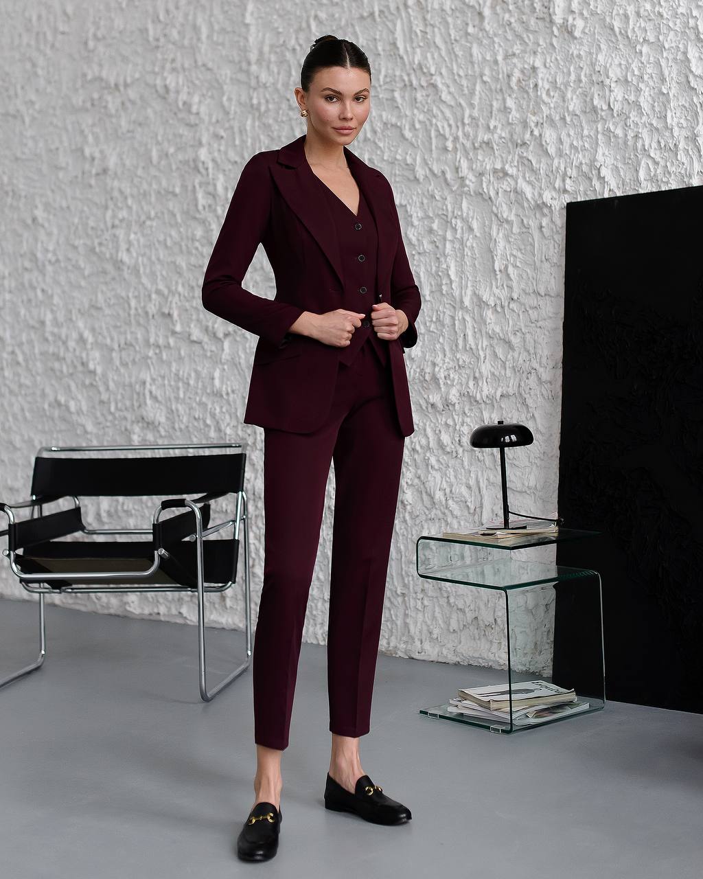 Woman in a burgundy suit standing in a modern room with a textured wall and black chair.