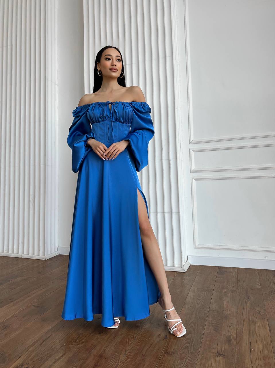 Woman wearing a blue off-shoulder dress with a slit, standing against a white paneled wall.