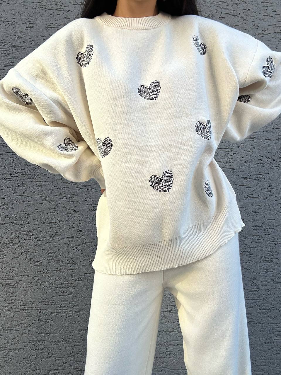 White sweater with black heart patterns worn by a person against a gray wall.