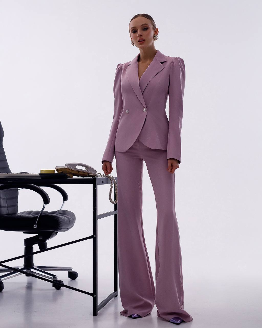 Woman in a pink suit standing in an office setting with a desk and chair.