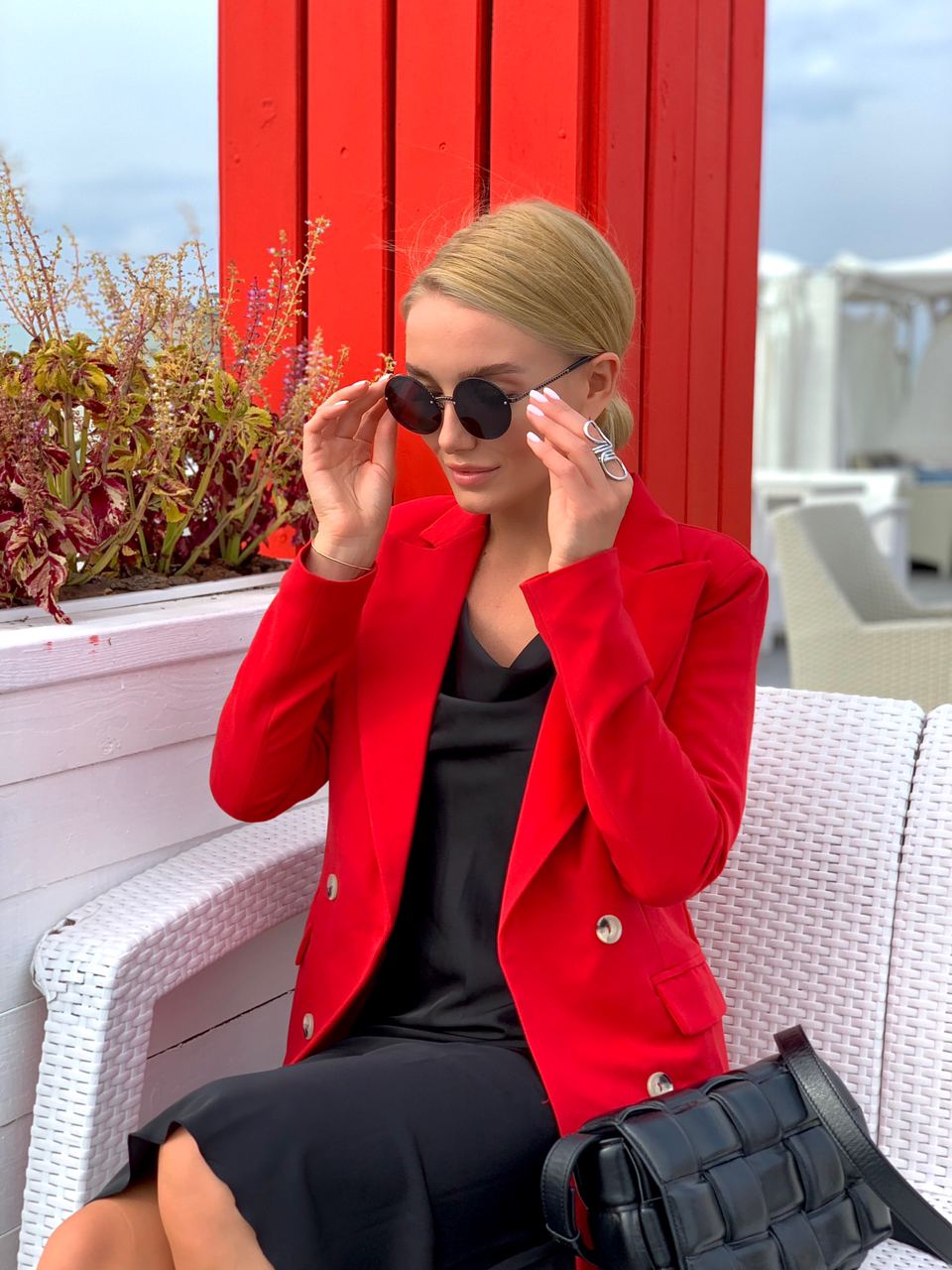 Woman in a red blazer and black dress sitting on a white bench with a red wall and plants in the background.