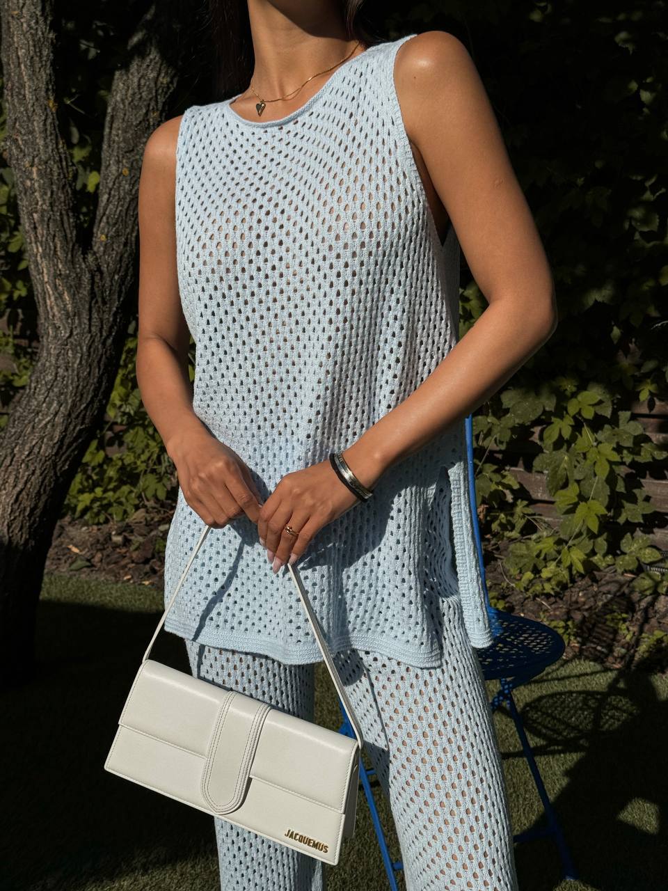 Person wearing a light blue sleeveless top and pants holding a white handbag outdoors.