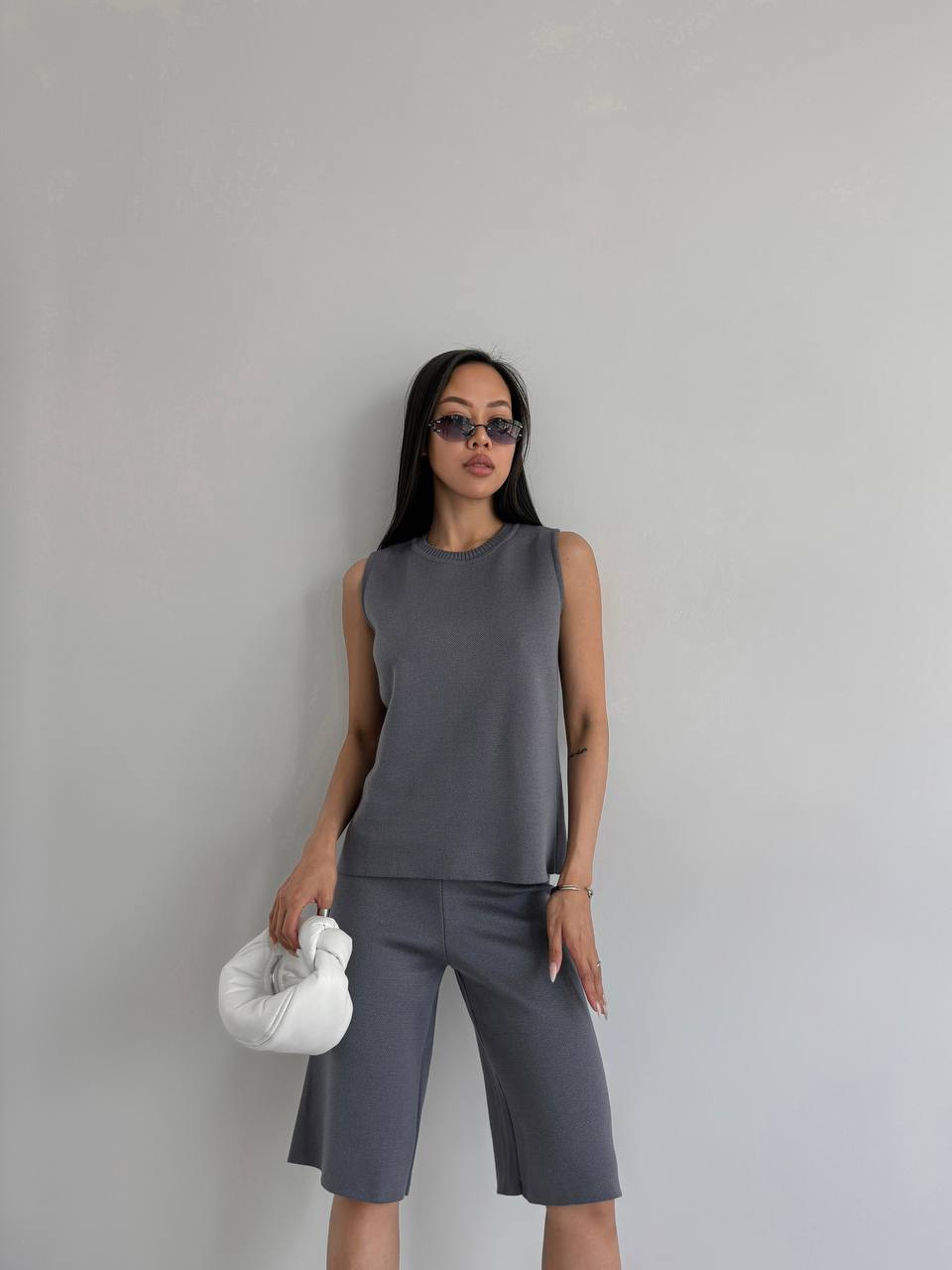 Woman wearing a gray sleeveless top and shorts holding a white bag against a plain background