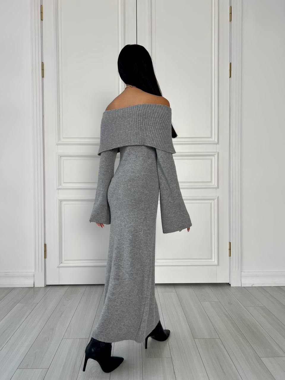 Person wearing a gray off-shoulder outfit standing in a room with white paneled walls.