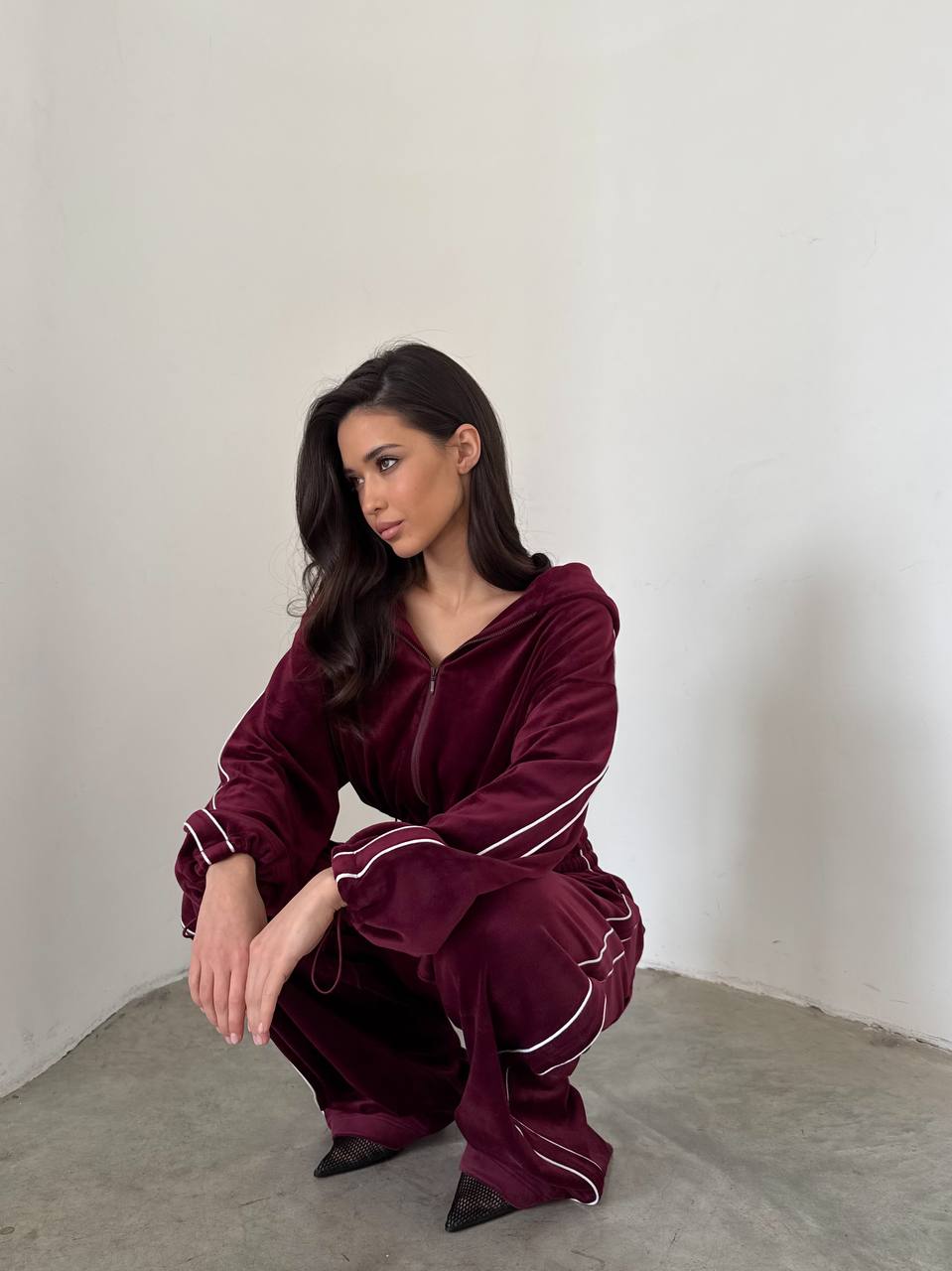 Woman wearing a burgundy velvet outfit with white piping, sitting on a gray floor against a white wall.