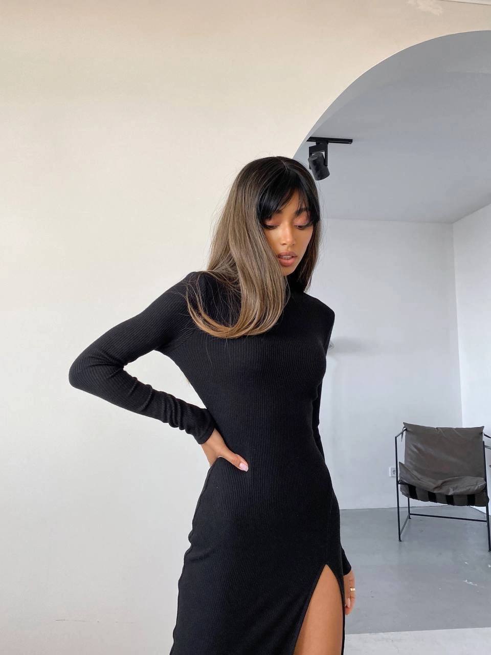 Woman wearing a black dress with a slit, standing in a minimalistic room.