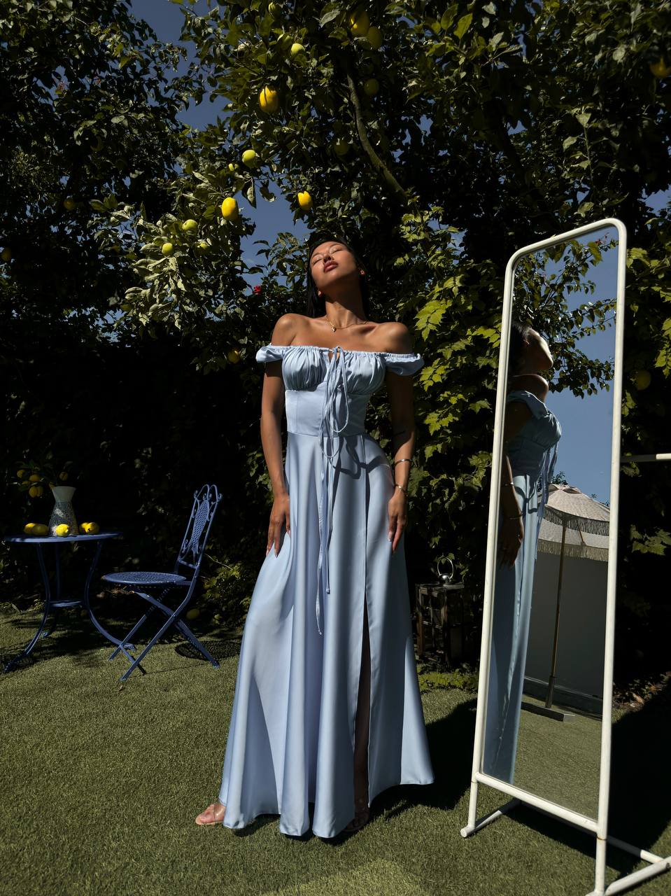 Woman in a light blue dress standing outdoors next to a mirror, with trees and a clear sky in the background.