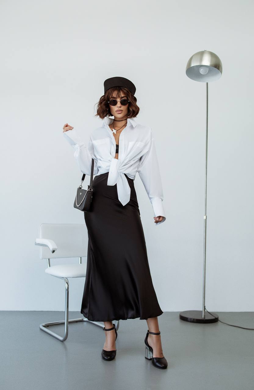 Woman in a white shirt and black skirt standing in a minimalistic room with a lamp and chair.