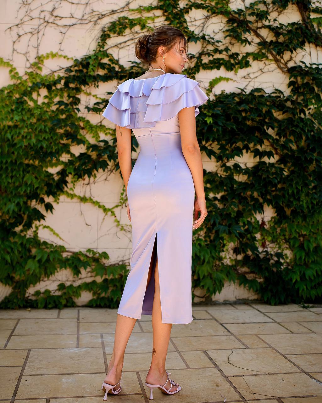 Woman wearing a light blue dress with ruffled details, standing against a wall with green ivy.