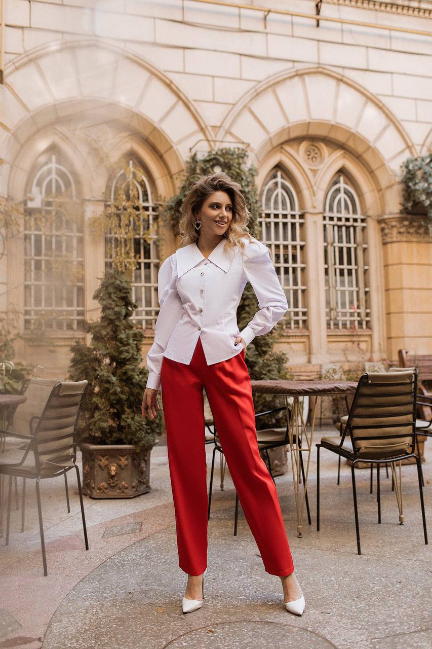 Woman in a white blouse and red pants standing in an outdoor setting with architectural elements.