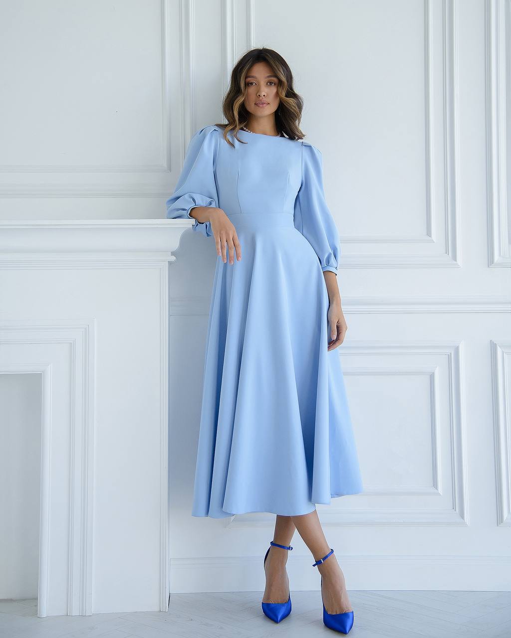 Woman wearing a light blue dress with long sleeves against a white paneled wall.