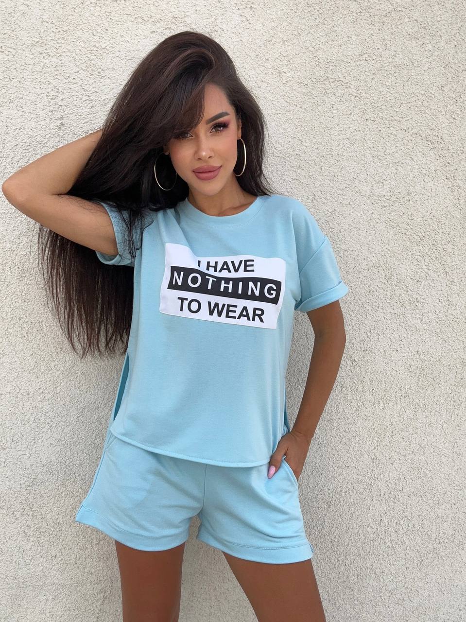 Woman wearing a light blue t-shirt with 'I have nothing to wear' text and matching shorts against a beige wall.