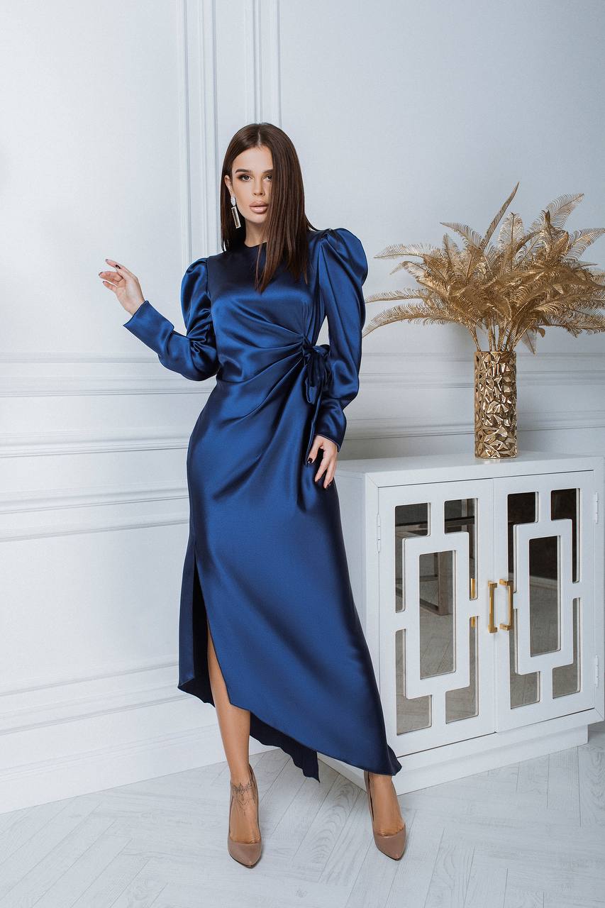 Woman in a blue dress standing in a room with a decorative cabinet and plant.