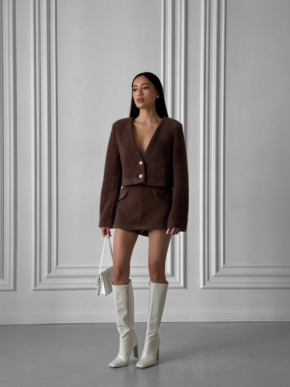 Woman wearing a brown outfit with white knee-high boots against a gray wall.