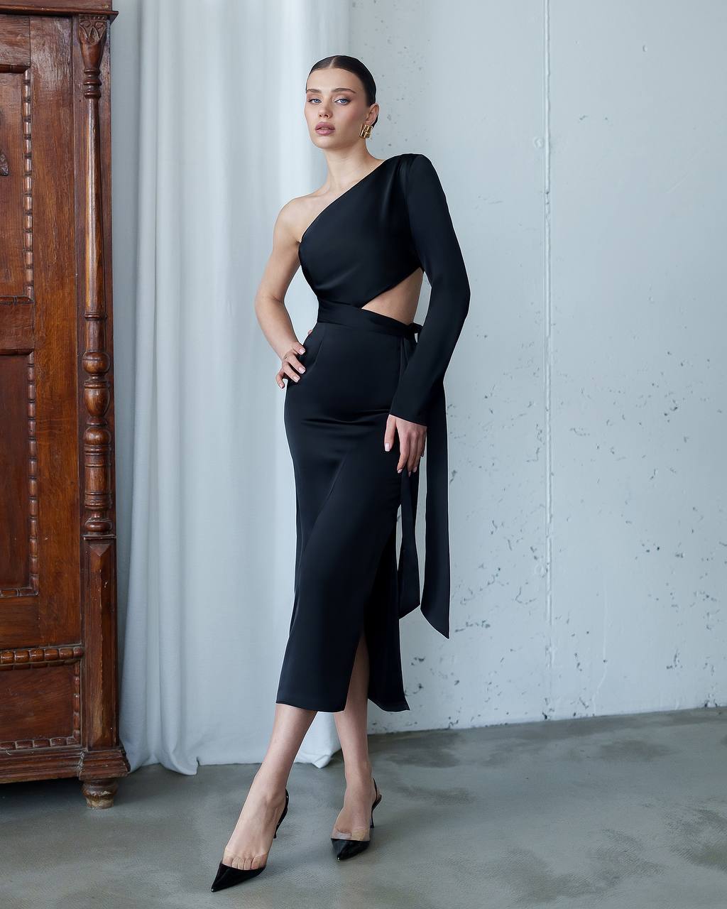 Woman in a black one-shoulder dress standing against a gray wall with a wooden cabinet.