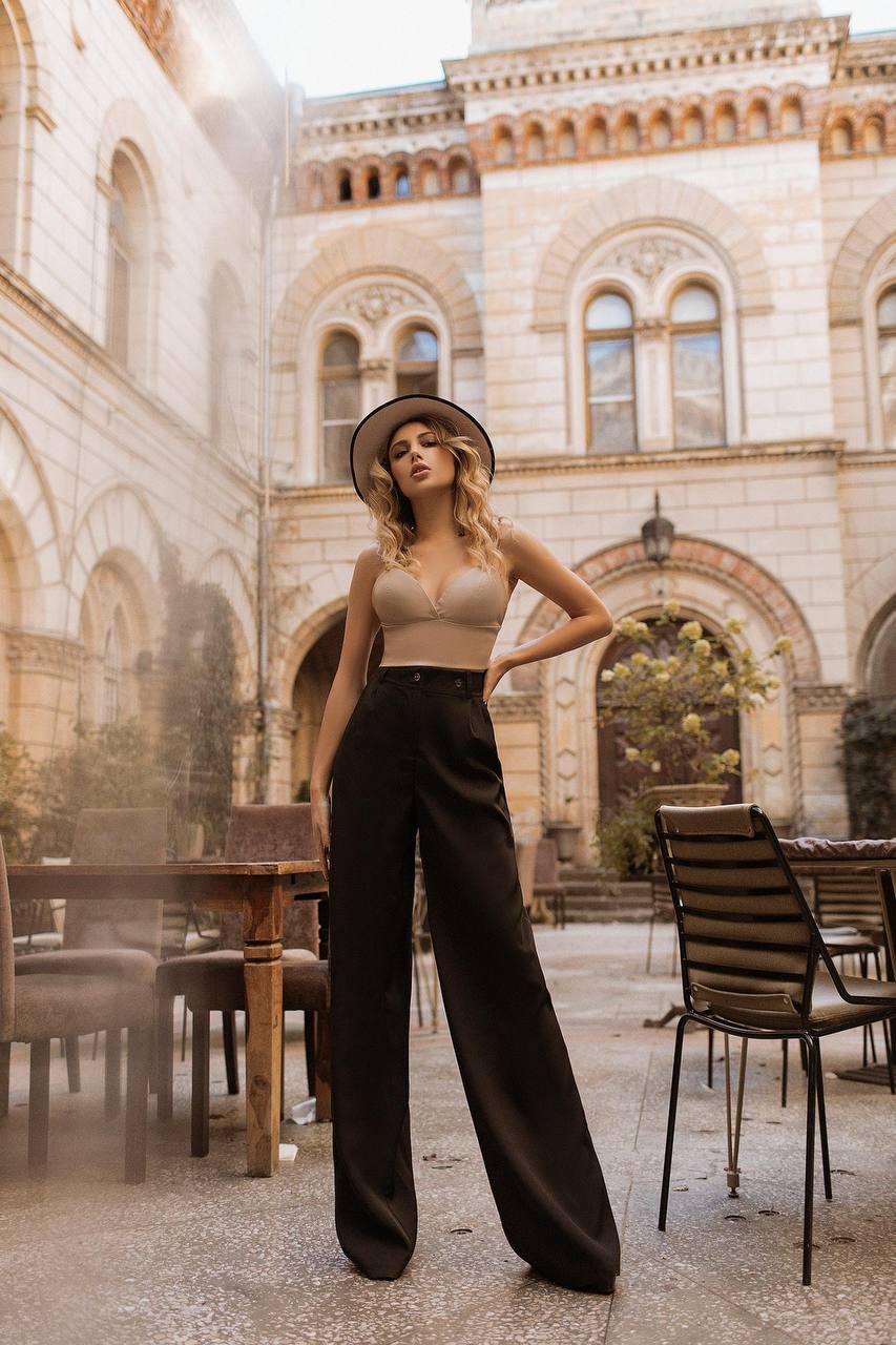 Woman posing in a stylish outfit with a hat in an outdoor setting with tables and chairs.