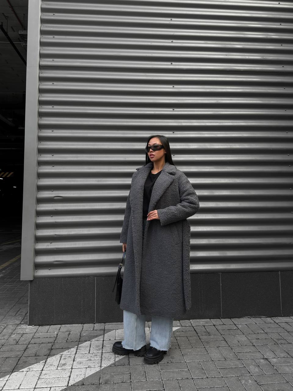 Person wearing a long gray coat standing in front of a corrugated metal wall.