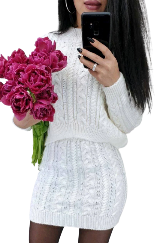 Person wearing a white cable knit sweater and skirt holding pink flowers and a phone.
