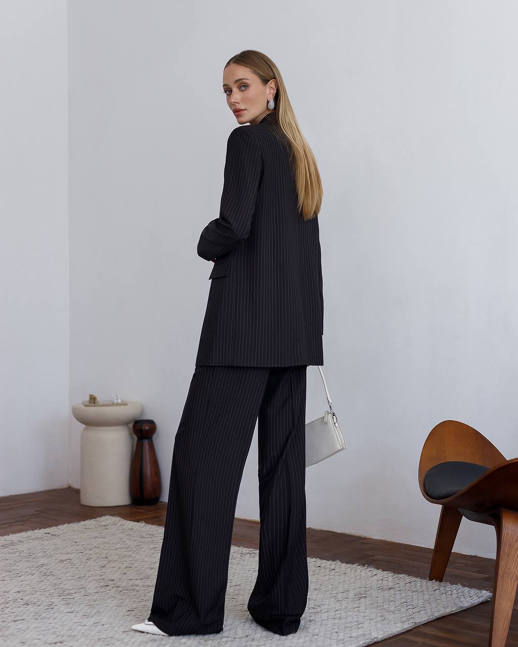 Woman in a black pinstripe suit standing in a minimalistic room with a white wall and wooden furniture.
