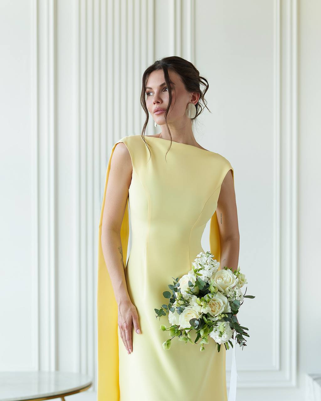 Woman in a yellow dress holding a bouquet of flowers against a white wall.